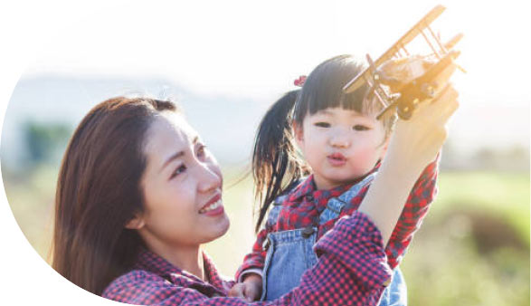 A mom hold a toddler girl (both Asian) outside. The mom holds a toy airplane.