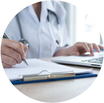 A light-skinned doctor is filling out paperwork on a clipboard with a pen while using a laptop.