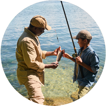 A father and son with brown skin stand in front of a lake as the son shows off a fish he caught.