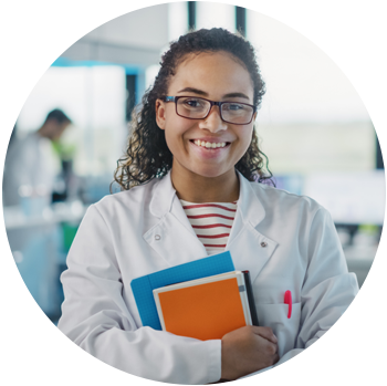 A young tan-skinned female scientist in a white lab coat holds books while smiling at the camera.