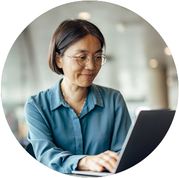 A middle-aged, medium-skin-toned Asian woman with glasses smiles and uses a laptop on a table.