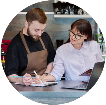 A white man and woman stand behind a counter at a cafe and look at a clipboard with paperwork. 