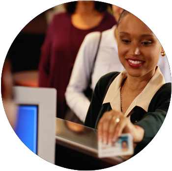 A young Black woman in business attire smiles and holds up her driver’s license to a DMV worker.