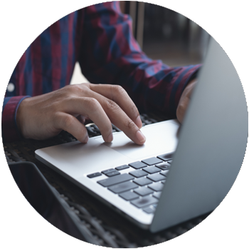 The hands of a man with tan skin and a checkered button-down shirt work on a laptop.