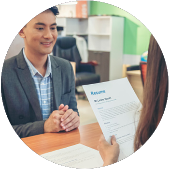 A young Asian man sits at a desk and smiles as a woman sits across from him and looks at his resume.