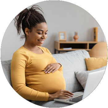 A young pregnant Black woman smiles and works on a laptop while sitting on a couch in a living room.