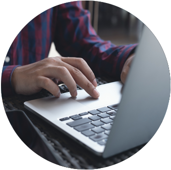 The hands of a man with tan skin and a checkered button-down shirt work on a laptop.