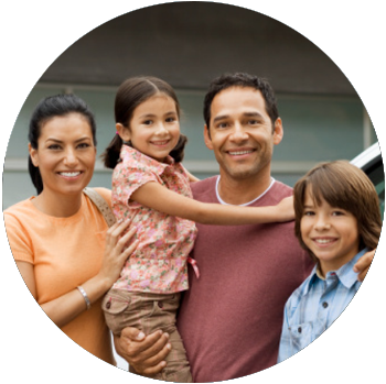 A tan-skinned family is smiling and standing outside of their home and next to their car.