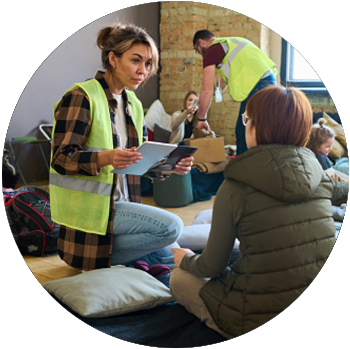 A tan female volunteer with a yellow safety vest holds a tablet and speaks to a person at a shelter.