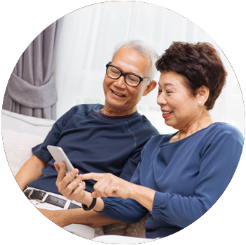 A senior-aged Asian couple smile while they sit on a couch and look at a mobile device.